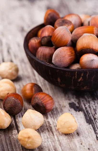 Avellanas enteras con cáscara en un cuenco de madera oscura, con frutos pelados y sin pelar sobre madera rústica